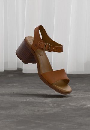 Sandale en cuir marron à bout ouvert avec talon bloc et bride de cheville à boucle, flottant au-dessus d'un sol gris avec des rideaux blancs en arrière-plan.