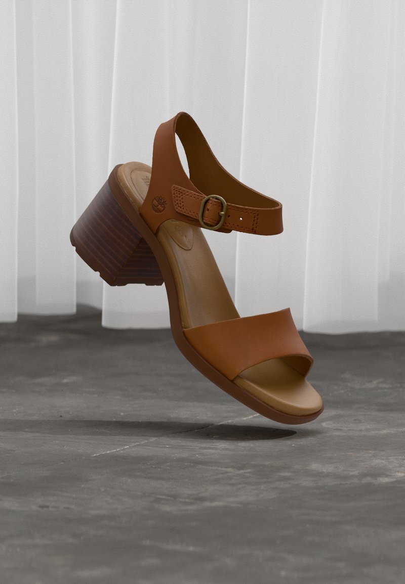 Καφέ δερμάτινο σανδάλι ανοιχτού δαχτύλου με χοντρό τακούνι και λουράκι στον αστράγαλο με πόρπη, αιωρούμενο πάνω από γκρίζο πάτωμα με λευκές κουρτίνες στο φόντο.