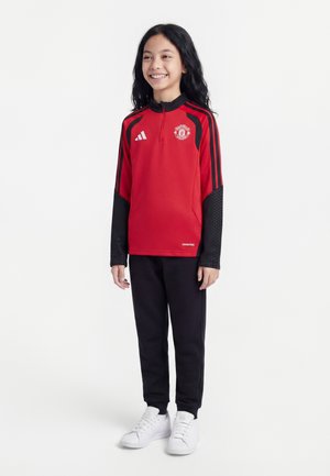 Uśmiechnięte dziecko z długimi czarnymi włosami, ubrane w czerwono-czarny pulower Manchester United, czarne spodnie i białe sneakersy, stojące na białym tle.