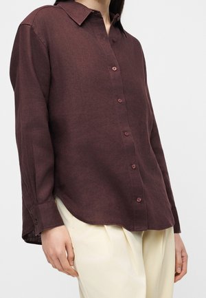 Femme portant une chemise ample à manches longues marron foncé boutonnée, associée à un pantalon beige clair sur un fond uni.