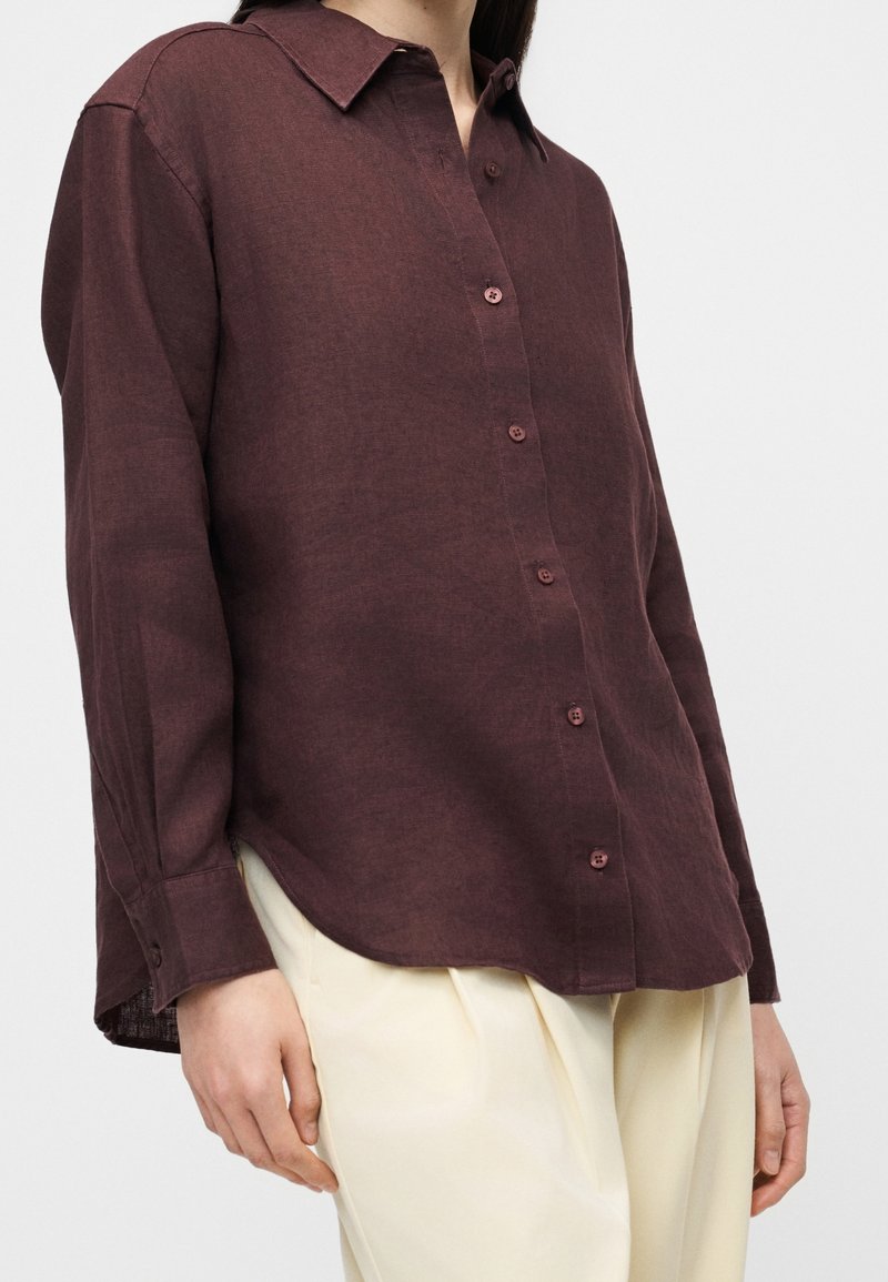 Femme portant une chemise ample à manches longues marron foncé boutonnée, associée à un pantalon beige clair sur un fond uni.