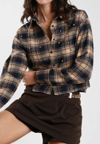 Donna che indossa una camicia a quadri marrone e nera con borchie, abbinata a una gonna marrone scuro con un dettaglio a cintura in catena dorata.