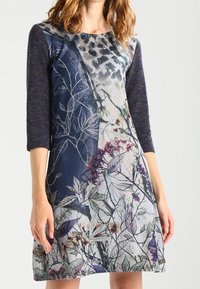 Robe à imprimé floral avec un corsage bleu foncé et des manches trois-quarts. Présente divers motifs de plantes en violet, vert et blanc.