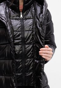 Fekete puff látózába, fényes befejezéssel, zippes elülső résszel, steppelt kialakítással és kapucnival. Két kéz tartja a kabát nyitását.