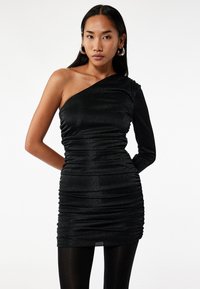 Robe noire à une épaule fabriquée en mat ériau brillant et texturé avec des détails froncés. S'ajuste parfaitement, se terminant au milieu de la cuisse, associée à des collants noirs.