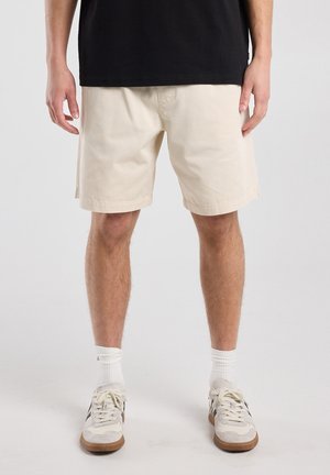 Homme portant un short beige aux genoux, des chaussettes blanches et des baskets blanches avec des détails noirs, debout devant un fond clair uni.