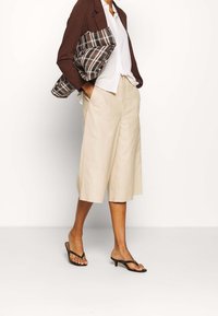 Beige culotter med vida ben, parat med en vit skjorta och en mörkbrun blazer. En rutig väska bärs under armen. Svarta flip-flops.