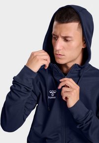 Navy zip-up hoodie in soft fabric features a front logo and adjustable drawstring hood. Model is tightening the zipper, showcasing the design.