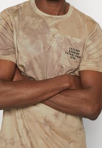 Béžové tričko s krátkým rukávem, které má strukturovaný maskáčový vzor, kulatý výstřih a kapsu na levé straně hrudi s černým textem "SAUCONY RUNNING ESTABLISHED 1898."