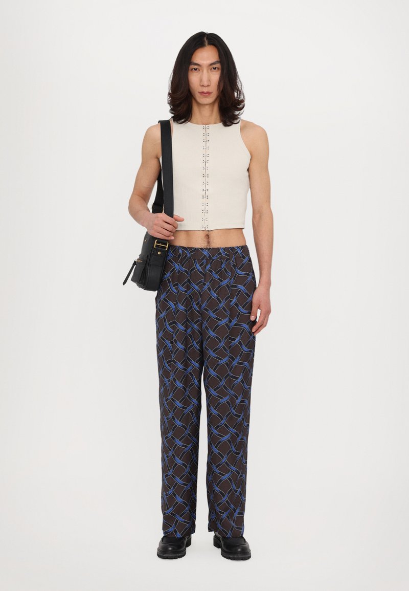 Top sans manches couleur beige clair, coupé court, avec des boutons sur le devant, associé à un pantalon ample à motifs sombres avec des lignes géométriques bleues.