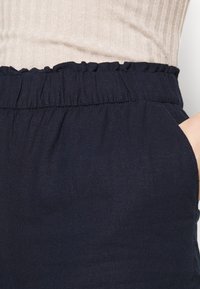 Námořnické modré kalhoty s elastickým pasem, se zdobeným okrajem. Materiál se zdá být lehký s hladkou texturou. Obsahují boční kapsy.