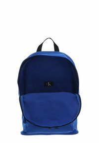 Sac à dos en tissu bleu de forme arrondie, avec une fermeture éclair noire et une étiquette logo à l'intérieur. Comprend un grand compartiment principal et une poche avant.