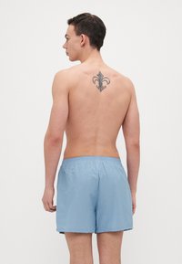 Shorts de bain bleu clair avec une taille élastique et des fentes latérales. L'arrière présente un motif de tatouage décoratif complexe.