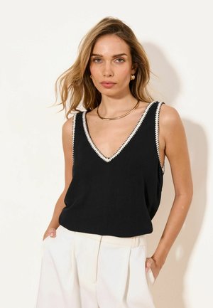 Femme aux cheveux ondulés et lâchés, portant un haut noir sans manches en V avec bordure blanche et un pantalon blanc taille haute, debout, mains dans les poches.