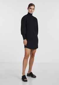 Robe en tricot noire avec un col montant, des manches bouffantes et des poignets côtelés. Associée à des chaussures à plateformes noires ouvertes à l'avant. Texture lisse.