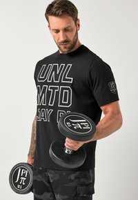 Černé sportovní tričko s bílým nápisem, černé maskáčové šortky a dvě černé činky s bílým brandingem, zdůrazňující zaměření na fitness.