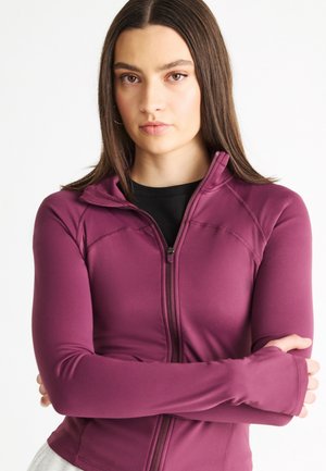 Ung kvinde med glat brunt hår iført en lyserød atletisk jakke med lynlås og tommelfingerhuller, armene krydsede, neutral udtryk, hvid baggrund.