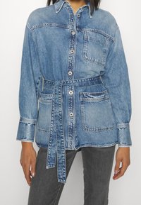 Jeansskjorta i ljusblå färg med knapprad fram, bröstficka, midjebälte och något oversizade ärmar. Slät yta, avslappnad design.