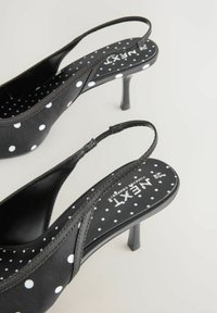 Pantofi cu toc înalt și barete negri, cu model cu buline, textură netedă și un toc subțire. Logo-ul brandului este vizibil pe branț.