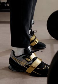 Scarpe da sollevamento pesi con un design nero e oro, realizzate in materiale sintetico, con inserti in rete e strap distintivi per una vestibilità sicura.