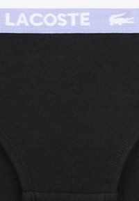 Juodi medvilniniai trumpikės su levandų spalvos liemeniu, kuriame matomas „LACOSTE“ ženklinimas ir logotipas. Lygus paviršius ir aptakus dizainas.