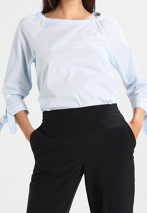 Light blue blouse with a wide neckline and gathered shoulders, featuring 3/4 sleeves with tie accents, paired with black trousers.