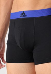 Črne prilegajoče se boksarice z modrim elastičnim pasom, na katerem je logo Adidas. Tkanina je gladka s podrobnostmi ploskega šiva.