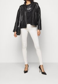Schwarze Leder-Übergroße Jacke über einem schwarzen Grafik-T-Shirt, kombiniert mit weißen Slim-Fit-Jeans und schwarzen spitzen Pumps. Kleine schwarze Tasche.