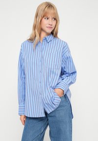 Világoskék, függőleges csíkos ing sötétkék és barna színben, mellkasi zsebbel és hosszú ujjakra hajtott mandzsettákkal.