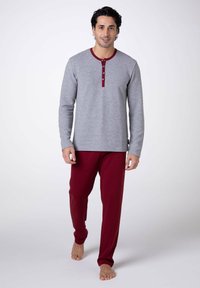 Maglia henley grigia a maniche lunghe con dettagli e bottoni rossi abbinata a pantaloni della tuta rossi. Il tessuto sembra morbido e la vestibilità è comoda. Piedi nudi.