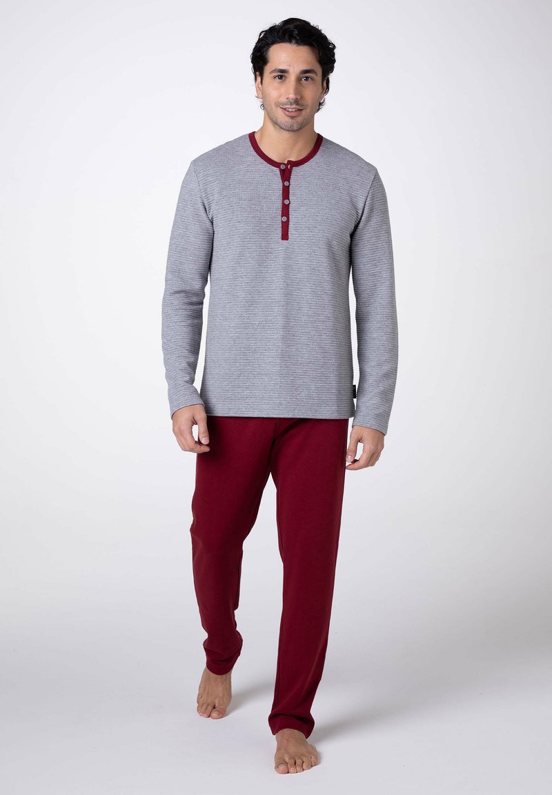 Maglia henley grigia a maniche lunghe con dettagli e bottoni rossi abbinata a pantaloni della tuta rossi. Il tessuto sembra morbido e la vestibilità è comoda. Piedi nudi.