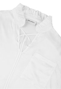 Hvit bluse med rysjekrage, snøring i halsen og venstre brystlomme. Glatt stofftekstur med subtil merkevarebrodering.