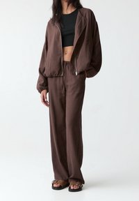 Hnědá lněná bunda se zipem vpředu, v kombinaci s volnými kalhotami ve stejné barvě. Černý crop top pod tím a ploché hnědé sandály s detaily na přezku.