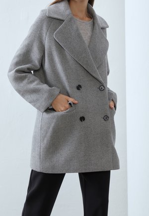 Femme portant un manteau croisé en chevrons gris avec des boutons noirs et les mains dans les poches, associé à un pantalon noir et une chemise grise.