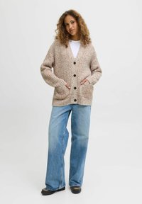 Beżowy sweter z dzianiny o teksturowanym wzorze, z przodem na guziki i dwiema dużymi kieszeniami, w połączeniu z jasnoniebieskimi spodniami typu wide-leg i czarnym obuwiem.