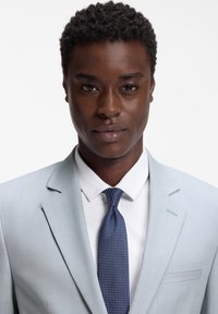 Άνδρας με κοντά σγουρά μαλλιά που φοράει ανοιχτό γκρι σακάκι, λευκό πουκάμισο και σκοτεινή μπλε γραβάτα με σχέδια, κοιτάζοντας μπροστά μπροστά σε λευκό φόντο.