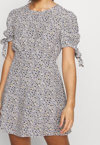 Lyhythihainen mekko pyöreällä pääntiellä, jossa on kukkakuosi mustassa, valkoisessa ja keltaisessa. Hihoissa olevat solmittavat yksityiskohdat tuovat lisäilmettä.