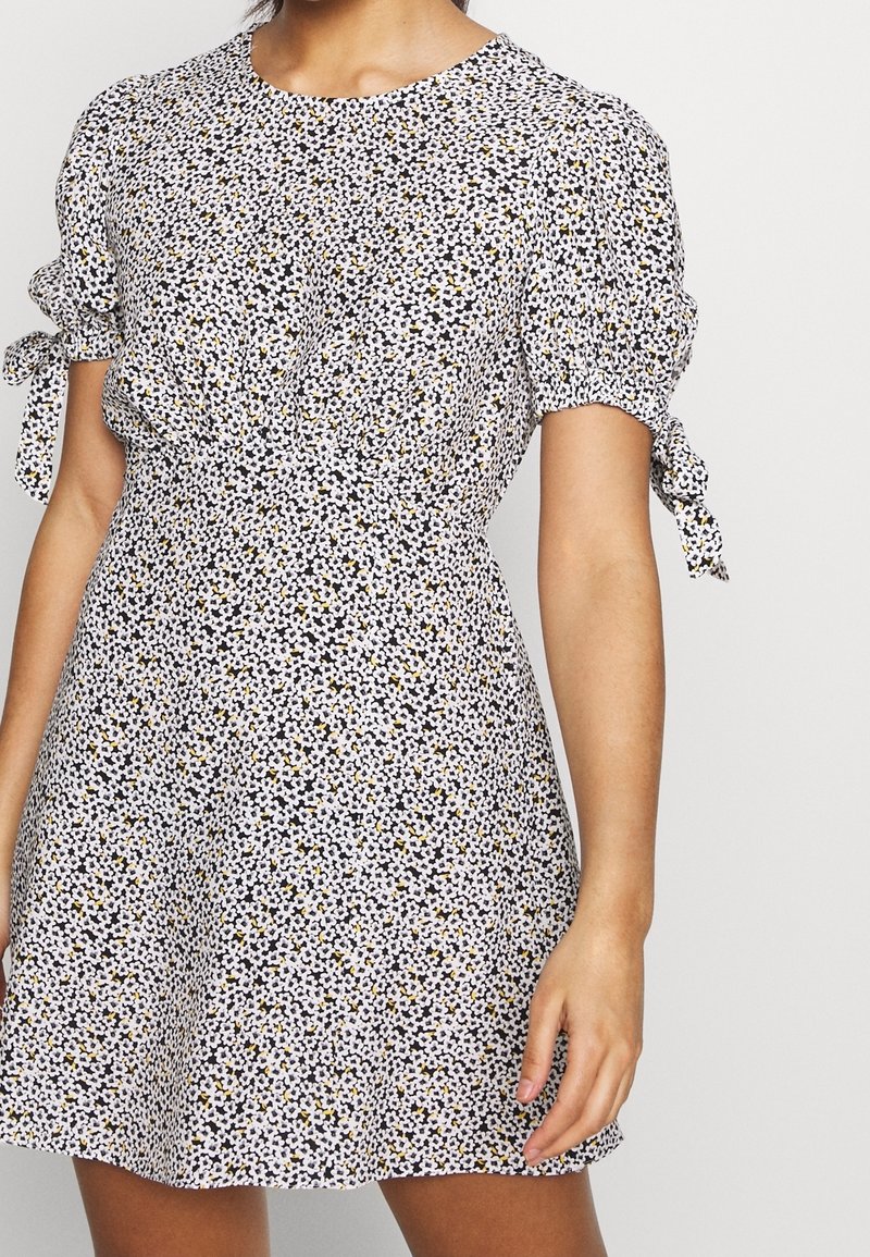 Lyhythihainen mekko pyöreällä pääntiellä, jossa on kukkakuosi mustassa, valkoisessa ja keltaisessa. Hihoissa olevat solmittavat yksityiskohdat tuovat lisäilmettä.