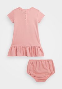 Lyhythihainen vaaleanpunainen mekko, jossa on röyhelöhelma ja takana nappikiinnitys, yhdistettynä samanvärisiin vaaleanpunaisiin shortseihin. Pehmeää puuvillakangasta.