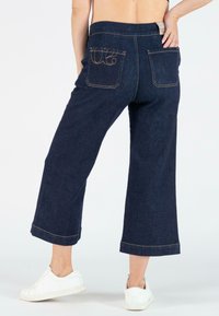 Donkerdenim wijde pijpen broek met een hoge taille, voorzien van achterzakken en contrasterende stiksels, gecombineerd met witte sneakers.