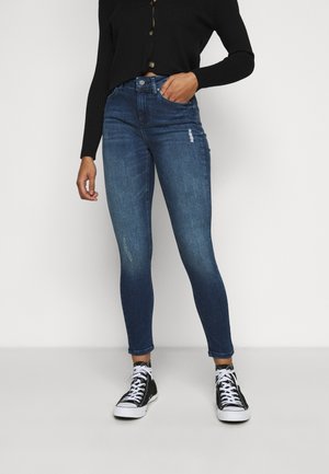 Personne portant un jean skinny bleu foncé avec de légères déchirures, un haut noir à boutons et des baskets montantes noires avec des lacets blancs.
