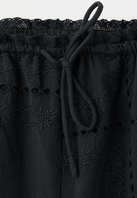 Tissu noir avec broderie florale délicate et motifs ajourés, doté d'une taille froncée avec cordon noué et bordure en dentelle en haut.