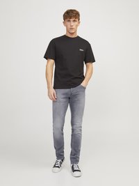Schwarzes kurzärmeliges T-Shirt mit einem kleinen weißen Logo, kombiniert mit hellgrauen Jeans und schwarzen Sneakers mit weißen Sohlen.