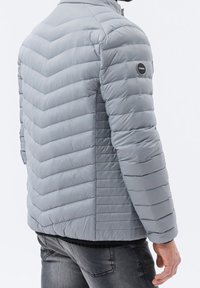 Szara kurtka puchowa z pikowaniem w jodełkę, wykonana z materiału syntetycznego, z wysokim kołnierzem i naszywką z logo na lewym rękawie.