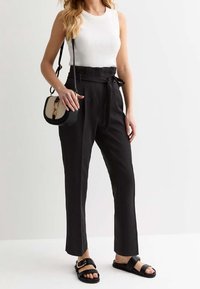 Zwarte high-waisted broek met een strikceintuur, gecombineerd met een aansluitend wit geribbeld tanktop en zwarte platte sandalen met een gespsluiting.