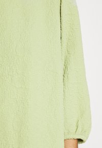 Haut vert clair avec une surface texturée, présenté dans une coupe ample et un tissu doux. Les manches sont légèrement bouffantes et finies par des poignets.