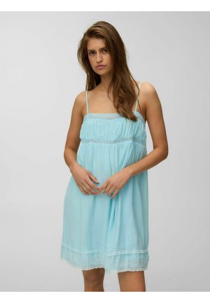 Giovane donna con lunghi capelli castani che indossa un vestito azzurro chiaro senza maniche con spalline sottili, in piedi davanti a uno sfondo grigio uniforme.