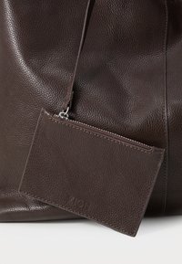 Hnědá kožená taška s hrubou texturou, která obsahuje odnímatelný držák na karty se zipovým uzávěrem a vyraženým logem.