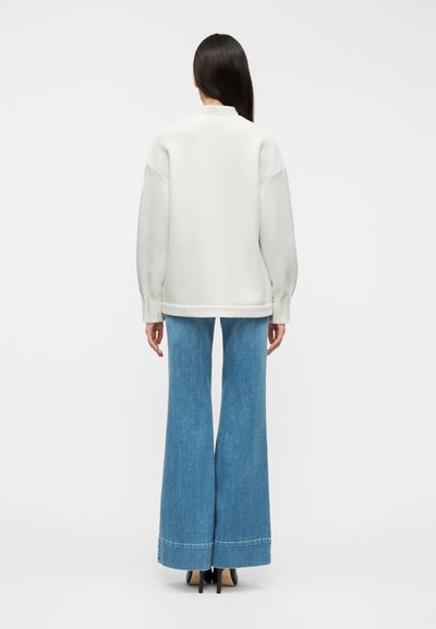 Λευκό φαρδιά πουλόβερ με κατεβασμένους ώμους και ριμπ μανσέτες, συνδυασμένο με ψηλόμεσα ανοιχτό μπλε τζιν με καμπάνα, ελαφρώς φθαρμένα στα τελειώματα.