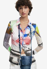Buntes Langarmhemd mit abstrakten Mustern in Pink, Grün und Blau; mit Kragen und Knopfleiste. Dazu eine Denim-Crossbody-Tasche.